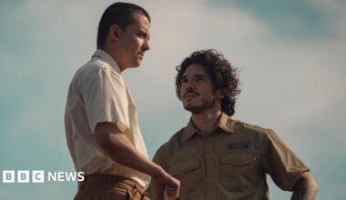 Asa Butterfield standing beside Fra Fee. Butterfield is wearing a short sleeve, cream coloured shirt. He has short brown hair. Fee is wearing a short sleeve, green shirt. He has short, curly brown hair.