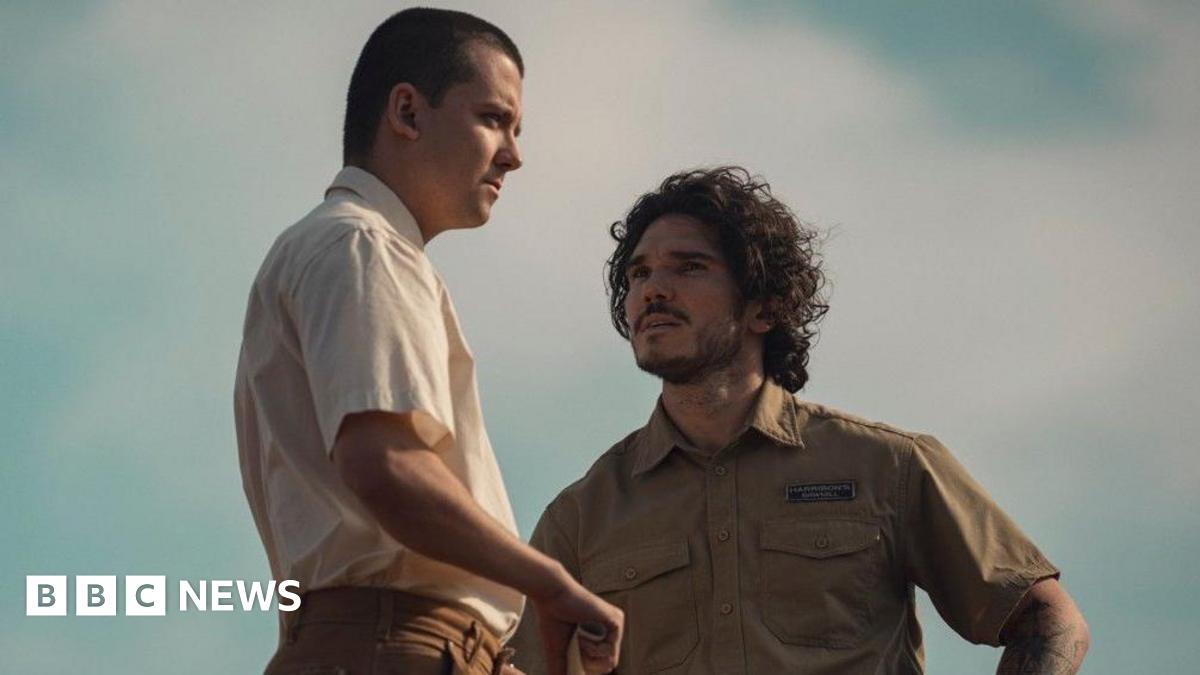 Asa Butterfield standing beside Fra Fee. Butterfield is wearing a short sleeve, cream coloured shirt. He has short brown hair. Fee is wearing a short sleeve, green shirt. He has short, curly brown hair.
