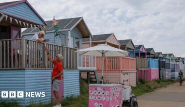 A row of beach huts in pastel blue and pink colours. A man stood next to an ice cream bike interacts with a woman on the terrace of a beach hut.