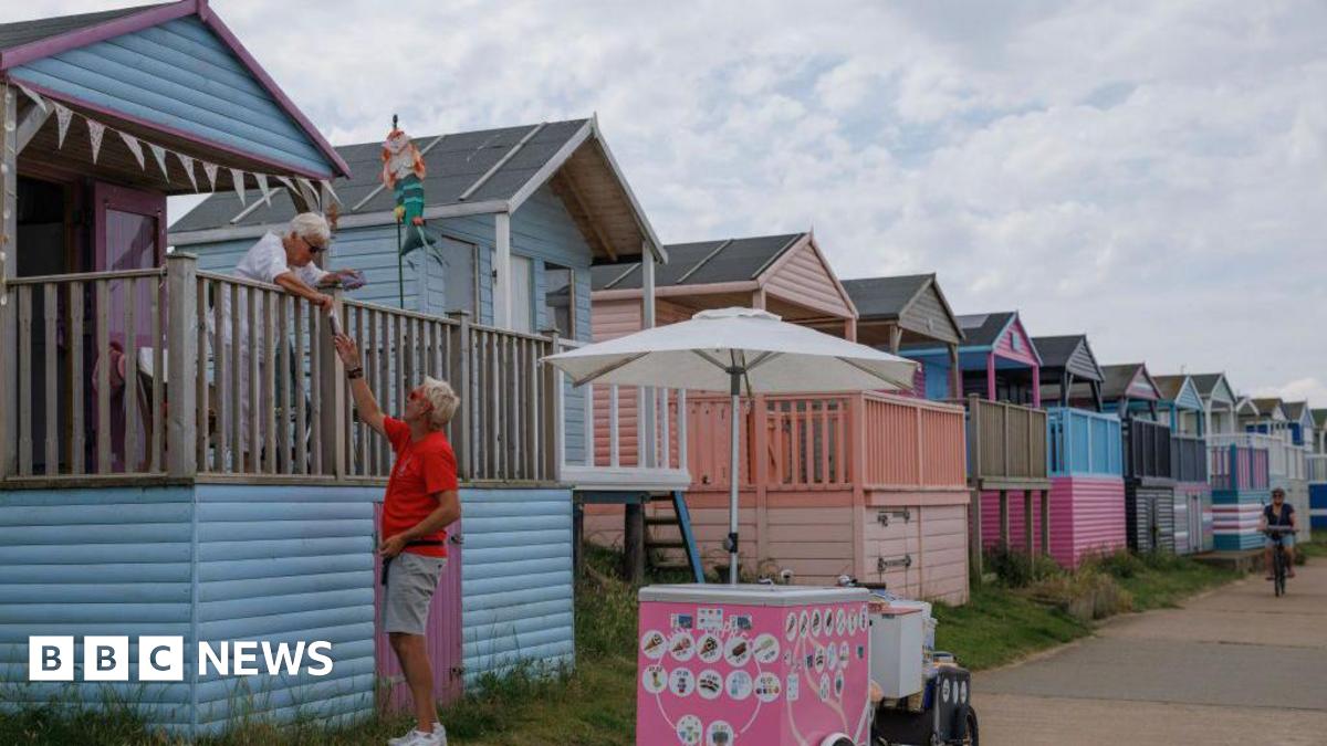 A row of beach huts in pastel blue and pink colours. A man stood next to an ice cream bike interacts with a woman on the terrace of a beach hut.