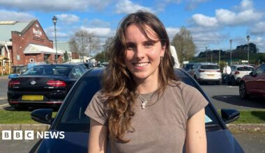 A female, wearing a grey top and necklace, smiling, standing in front of a dark coloured car, in a car park. Behind her on the right is a maroon car and on the left a black car with a blurred out number plate.