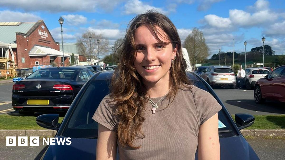 A female, wearing a grey top and necklace, smiling, standing in front of a dark coloured car, in a car park. Behind her on the right is a maroon car and on the left a black car with a blurred out number plate.