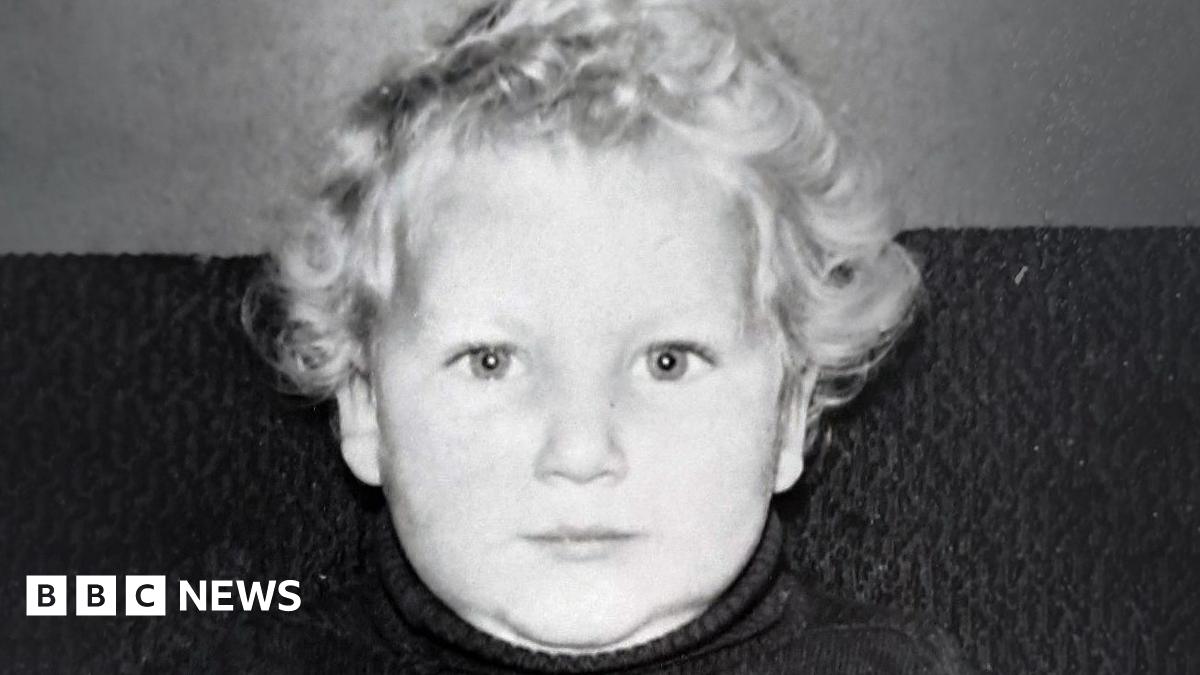 A black and white image of a young boy with blond hair, looking into the  camera with a smile