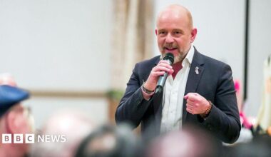 Steve Hilton speaks at a campaign event. He holds a microphone, and wears a black blazer with a pin in the shape of California.