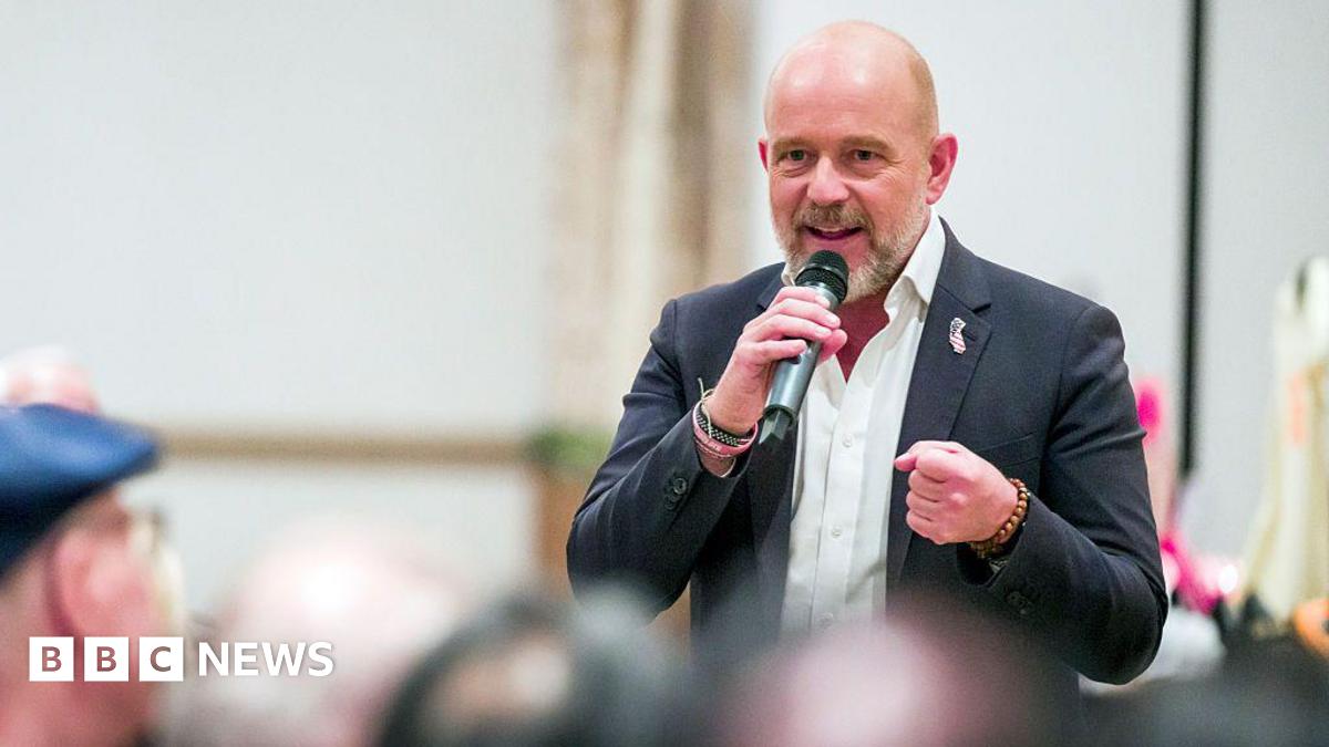 Steve Hilton speaks at a campaign event. He holds a microphone, and wears a black blazer with a pin in the shape of California.