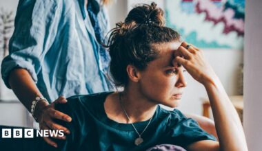 A sad woman sitting on a sofa, a person behind them has placed their hand on her shoulder in empathy.