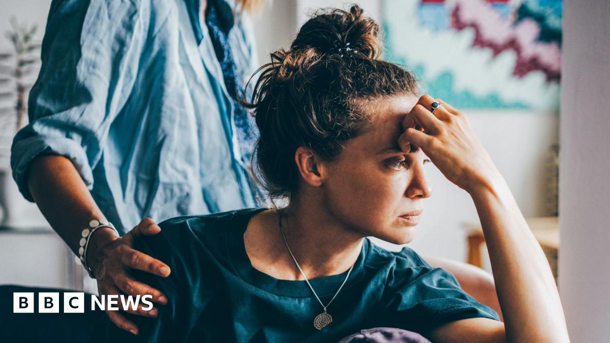 A sad woman sitting on a sofa, a person behind them has placed their hand on her shoulder in empathy.