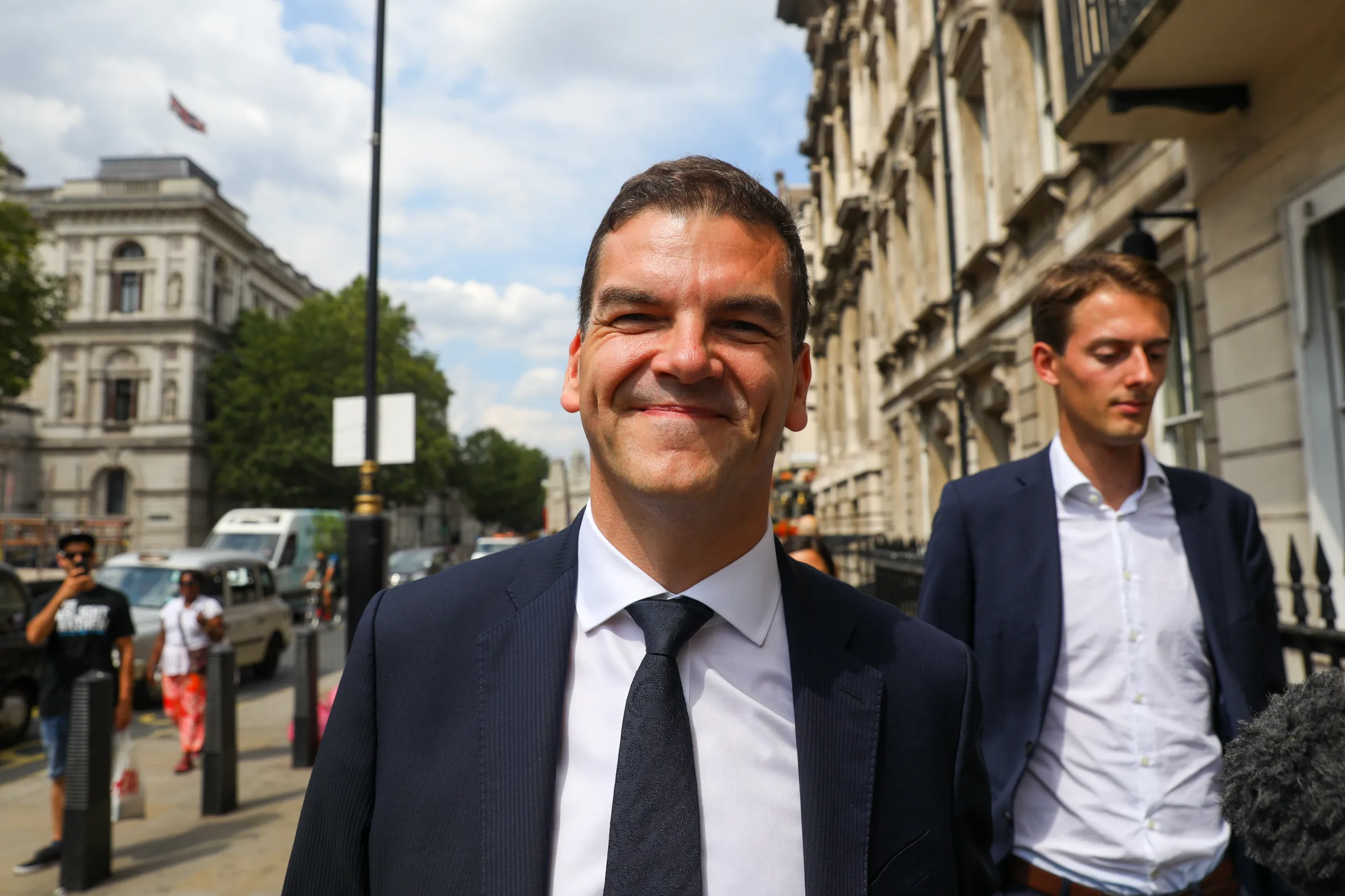 Olly Robbins, Brexit adviser to Prime Minister Theresa May, smiling and wearing a suit and tie, with another man and a city street in the background.