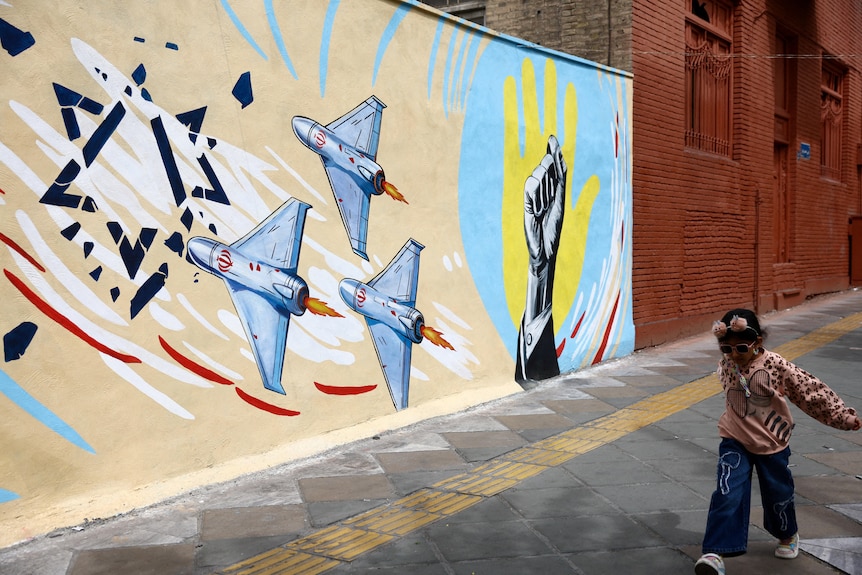 A young girl skips in front of a wall with a painting of warplanes attacking the Star of David with a fist next to them.