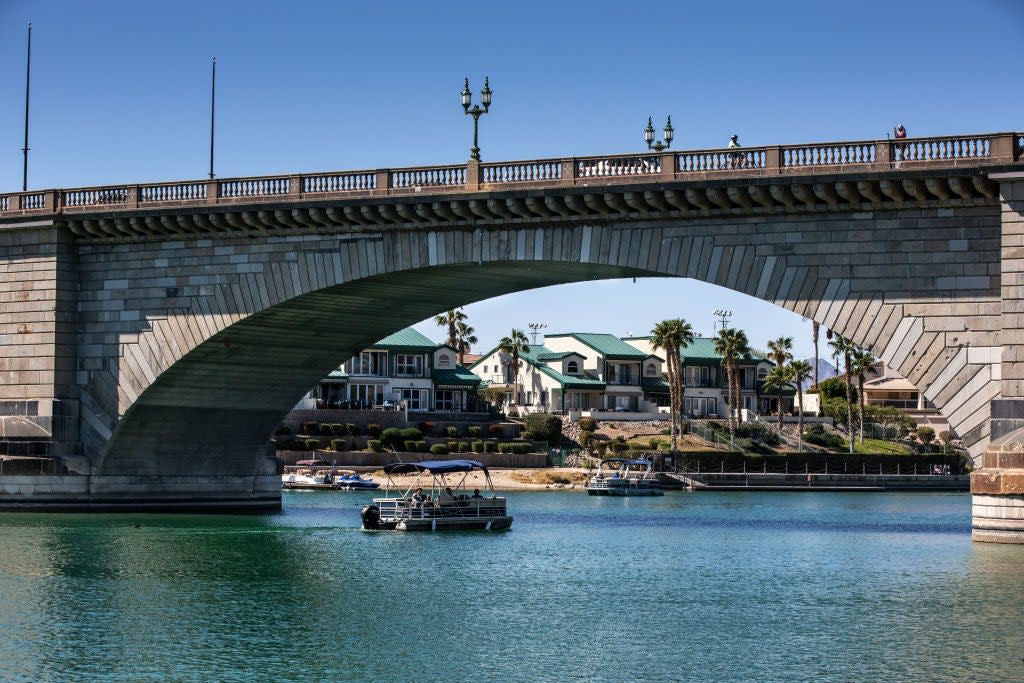 Stone bridge over a river with a pontoon boat beneath; houses and palm trees are visible in the background
