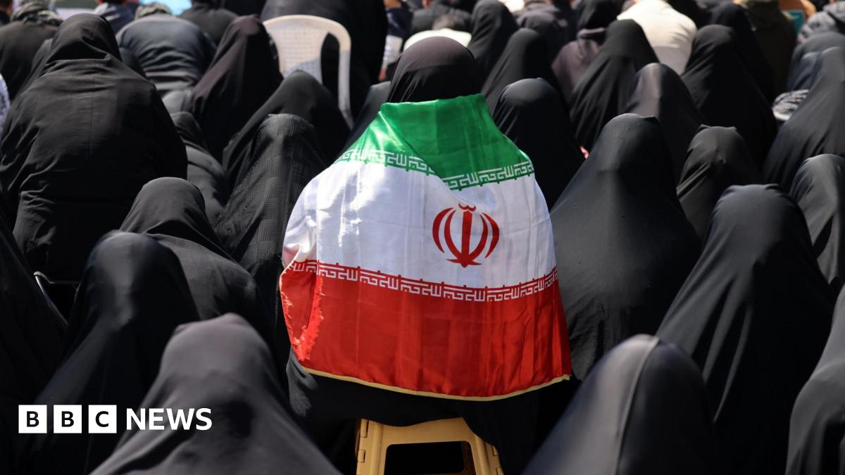 An Iranian woman wears a black hijab and chador and has a national flag draped over her shoulders as government supporters gather at Enghelab Square following the US-Iran ceasefire announcement, in Tehran, Iran (8 April 2026)