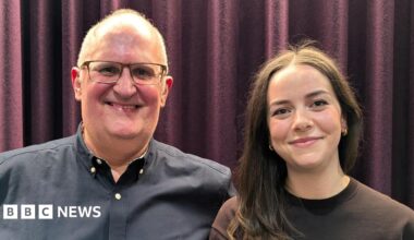 A smiling woman with blonde hair wearing a London Ambulance polo shirt with NHS and ambulance service logos on the front and epaulettes on the shoulders. An ambulance is in the background.