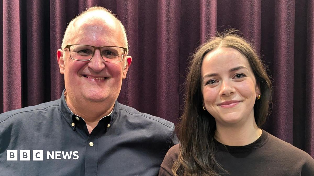 A smiling woman with blonde hair wearing a London Ambulance polo shirt with NHS and ambulance service logos on the front and epaulettes on the shoulders. An ambulance is in the background.