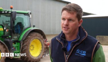 Ashley Jones wears a gilet over a green top and stands side on to the camera talking to an out of shot journalist with a serious expression. Behind him to the left is a large green tractor with yellow wheels.