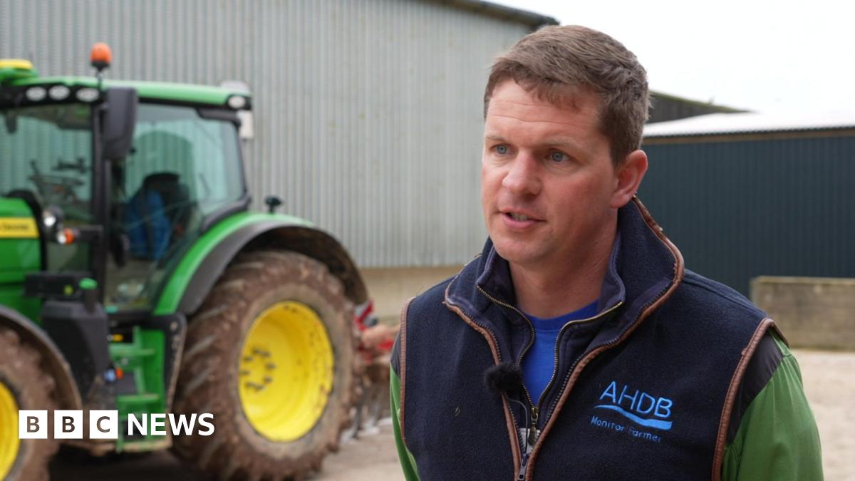 Ashley Jones wears a gilet over a green top and stands side on to the camera talking to an out of shot journalist with a serious expression. Behind him to the left is a large green tractor with yellow wheels.