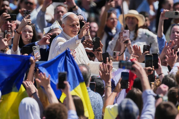 A man in white robes amid an outdoor crowd smiles on a sunny day. people wave blue and yellow ukrainian flags.