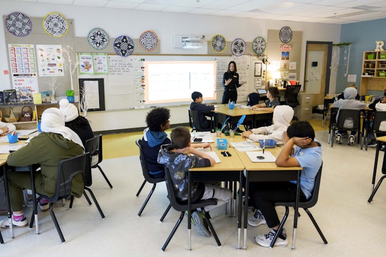 A teacher seen in a classroom with children at desks.