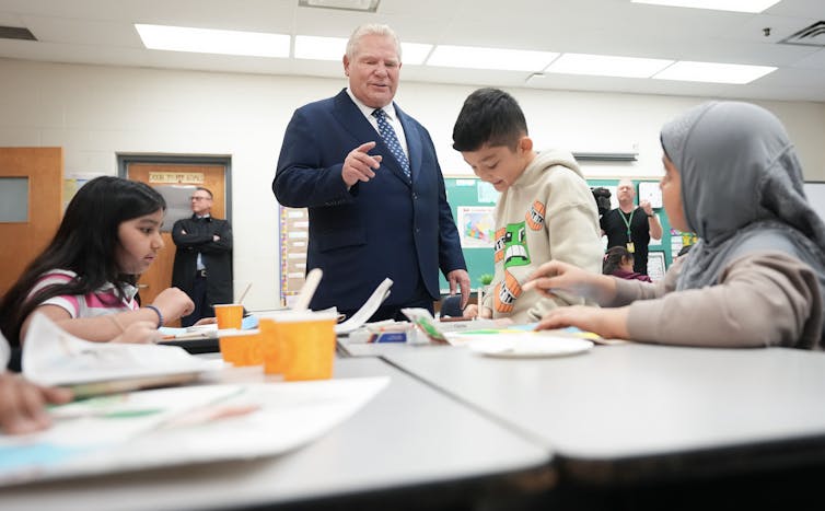 A man in a suit standing in a classroom next to students working at desks.