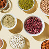 Various foods in bowls on a beige background. The foods include sources of dietary fiber such as mung beans, chickpeas, lentils, kidney beans, almonds and hazelnuts.