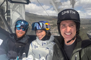 Two men and a woman smile while riding a ski lift up the slopes.