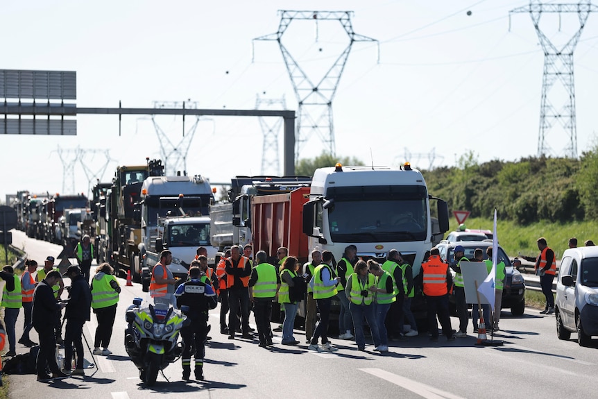 Crowds of workers stand on the road as a long line of traffic builds behind them during a protest.