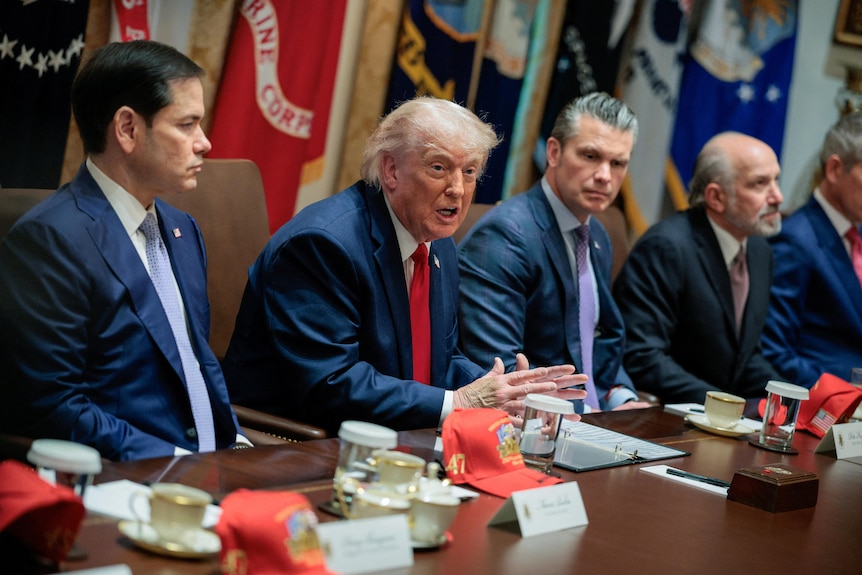An eldely man with thinning hair speaks at a meeting table in the White House.