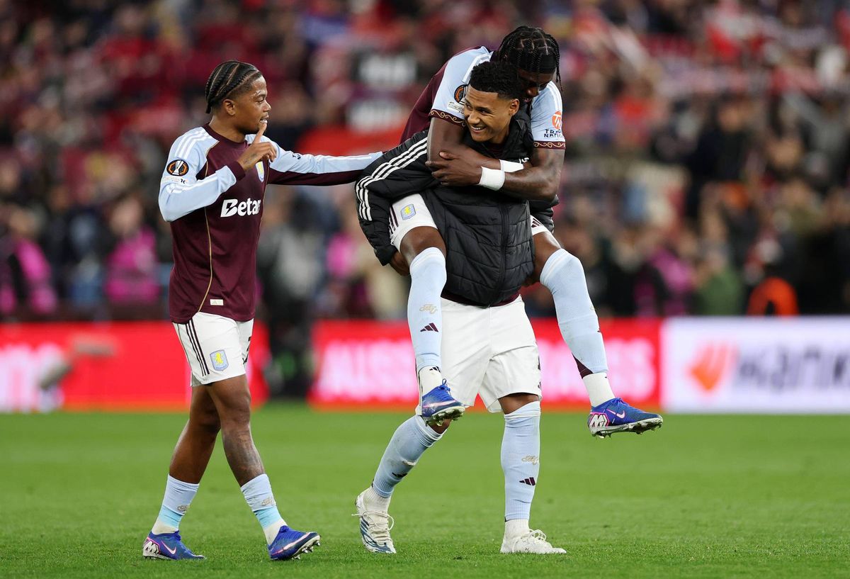 Two athletes, dressed in athletic uniforms, celebrate on a field in front of a crowd. One athlete is lifted into the air by another while both embrace each other.