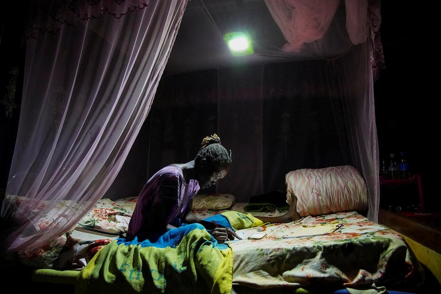 A girl studies cross-legged on her bed, her canopy bed lit by a single solar light beaming down from the ceiling.