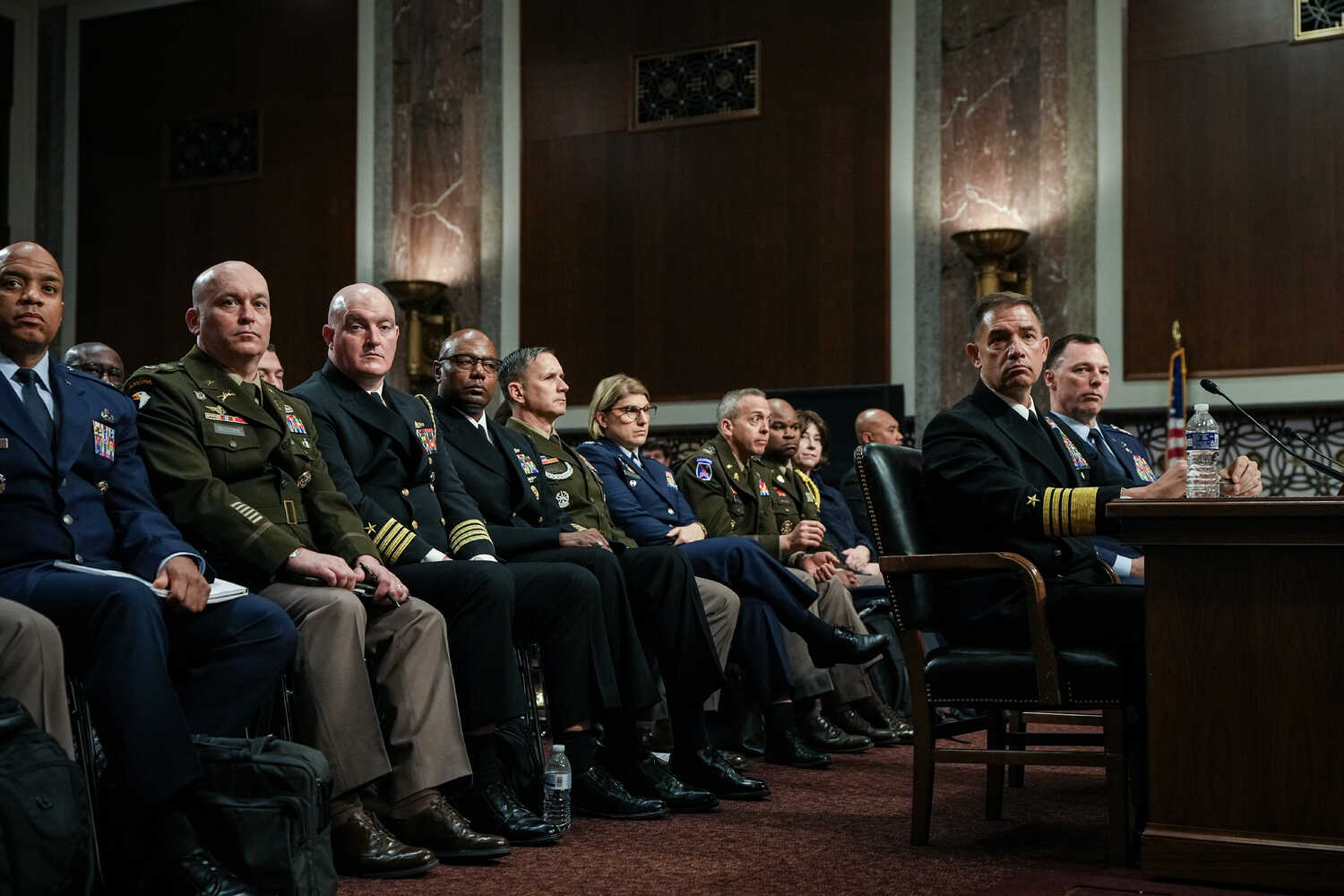 Mr. Cooper and Mr. Anderson sit at a desk while various members of the U.S. military sit behind them.