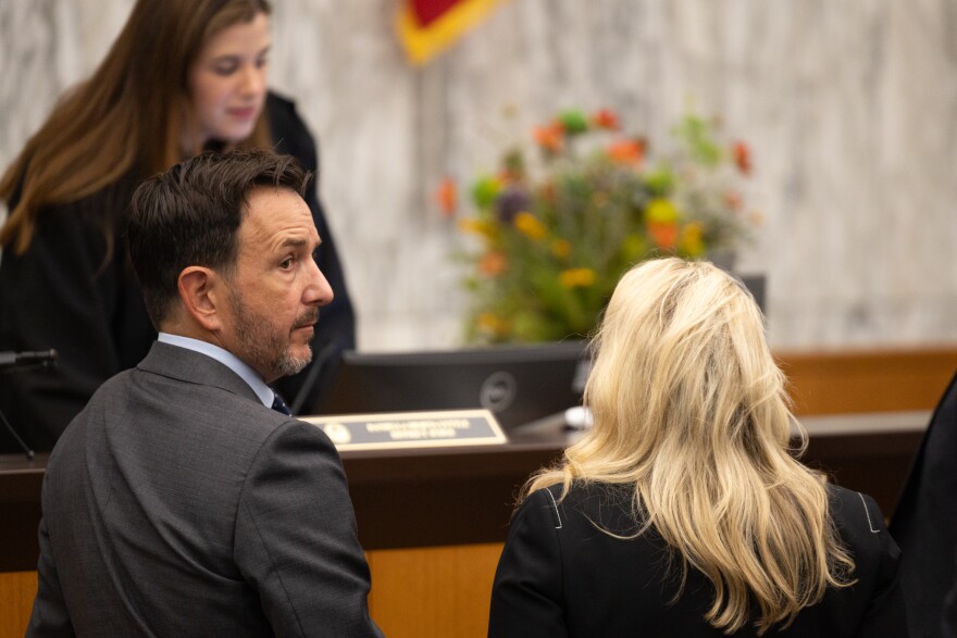 A man in a dark suit in a court room turns his head to glance behind him.