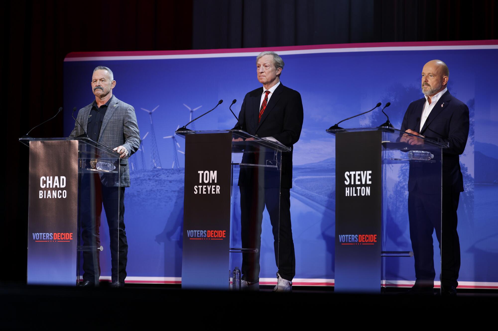 Chad Bianco, left, Tom Steyer and Steve Hilton stand behind podiums and speak into microphones.