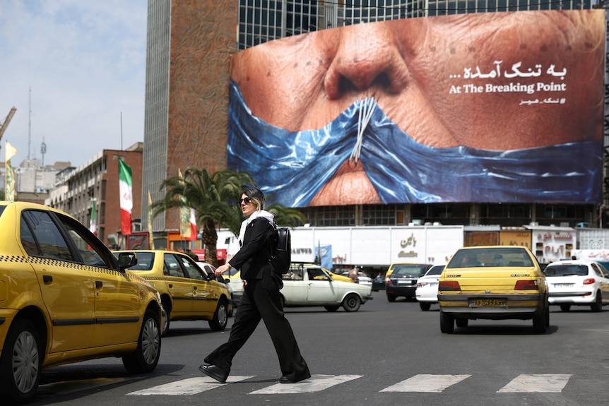 A woman walks across a Zebra crossing near some Taxis. In the background is a billboard depicting Trump with his mouth shut.
