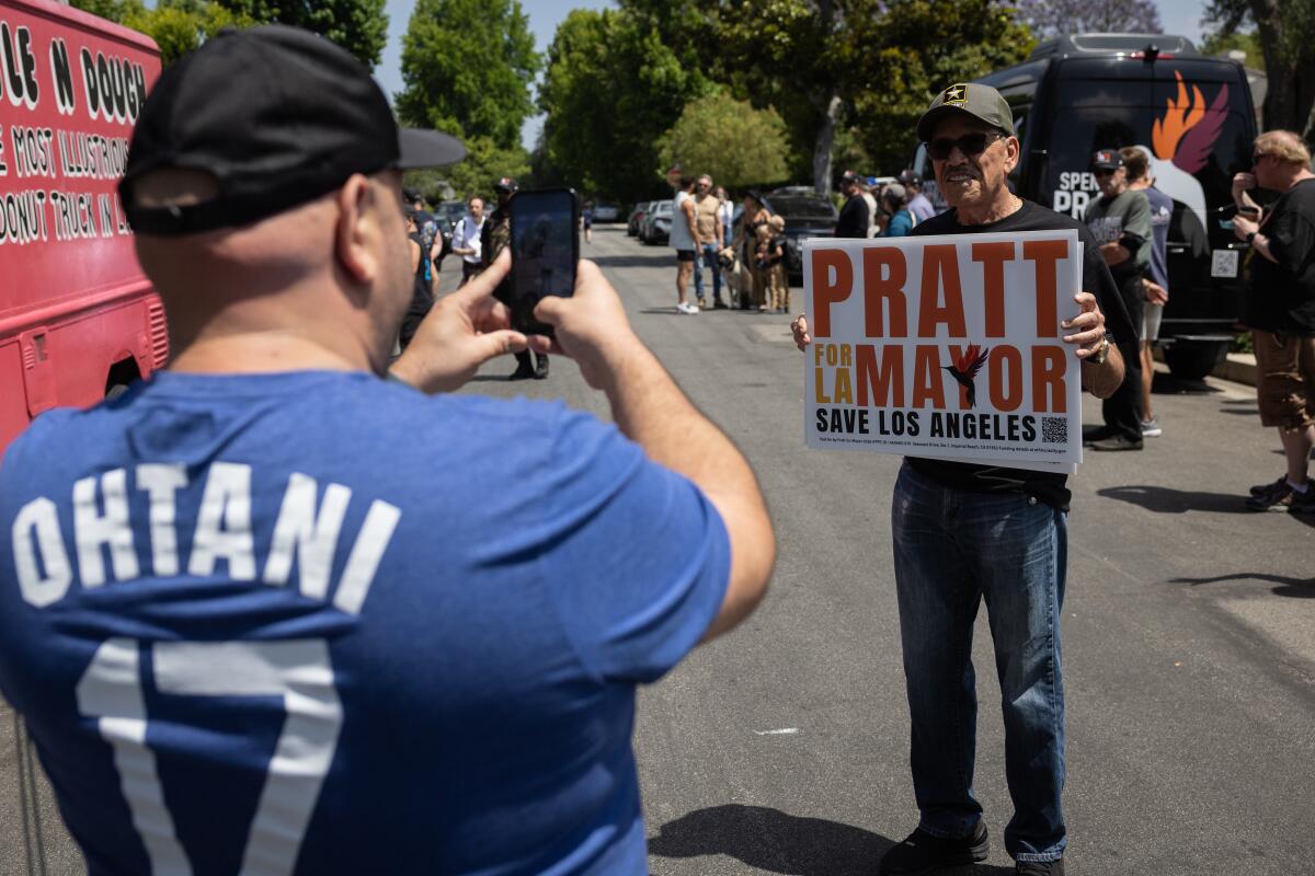 A man photographs his father holding a "Pratt for L.A. Mayor" sign in the street