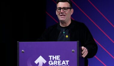 Andy Burnham delivers speech, standing at podium, emphasizing key points, wearing a suit, addressing an engaged audience