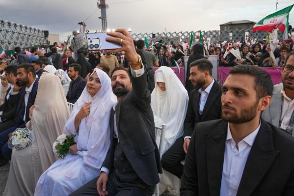 A groom takes a selfie as couples sit during a mass wedding ceremony for participants in the "Janfada" ("Sacrifice for Iran") pro-government campaign in Tehran, Iran, Monday, May 18, 2026.(AP Photo/Vahid Salemi)
