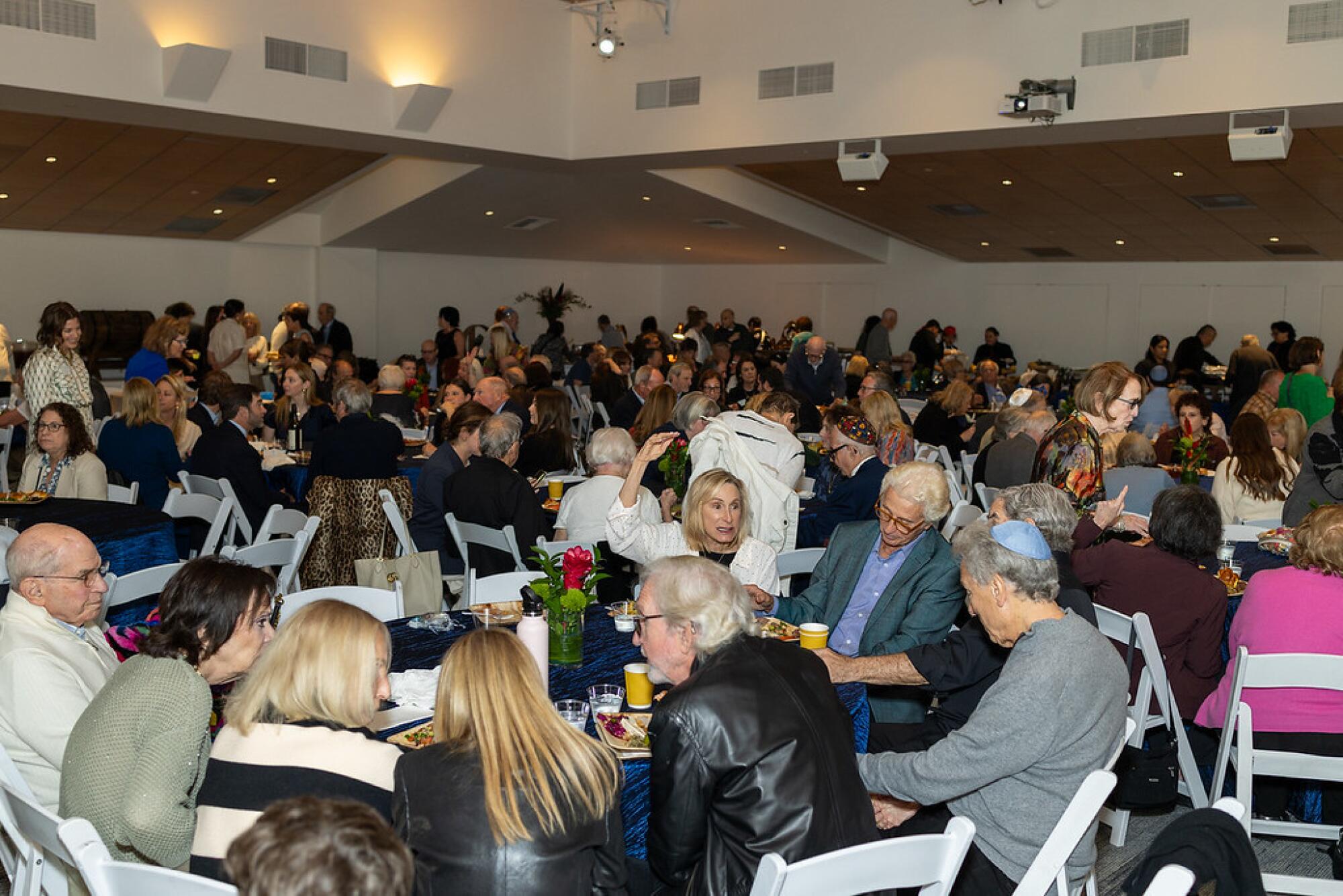 A temple hall is filled will people sitting at round tables with blue tablecloths.