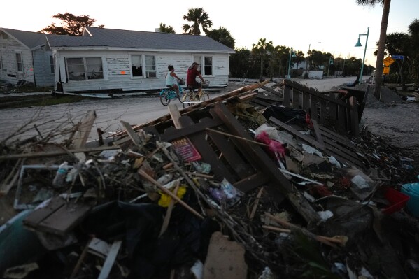 People bike past damaged homes and debris left by Hurricane Milton, on the sand-coated main road of southern Manasota Key, already cleared of feet of sand, in Englewood, Fla., Oct. 13, 2024. (AP Photo/Rebecca Blackwell, File)