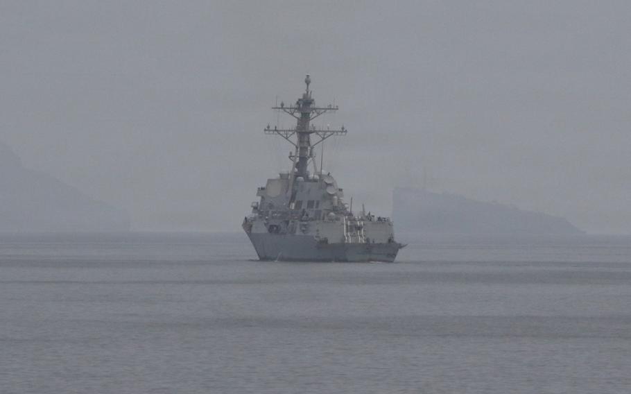A U.S. military warship is seen from behind and at a distance as it transits open ocean through hazy skies, with the horizon line in the background.