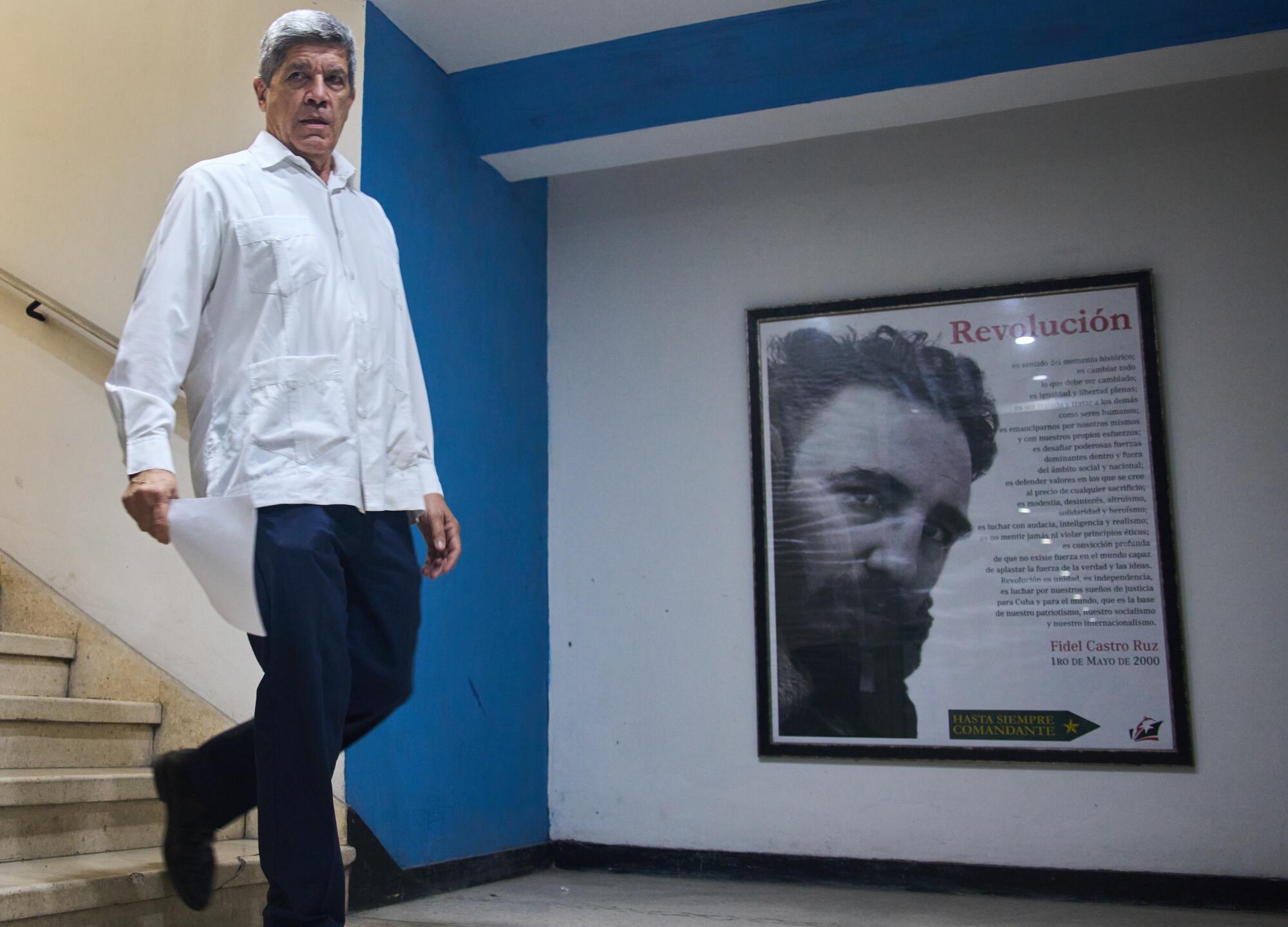 A man in a white shirt and dark pants descends a stairway, near a wall with a framed image of a dark-bearded man