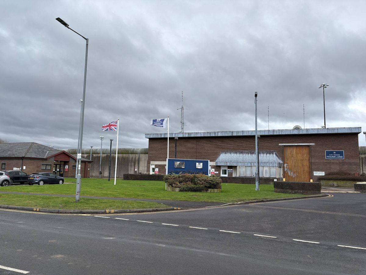 An image depicts an intersection with a building situated in the background. The building features multiple flags and a grassy area in front. Street lights and a road with white markings are visible. The sky is overcast.
