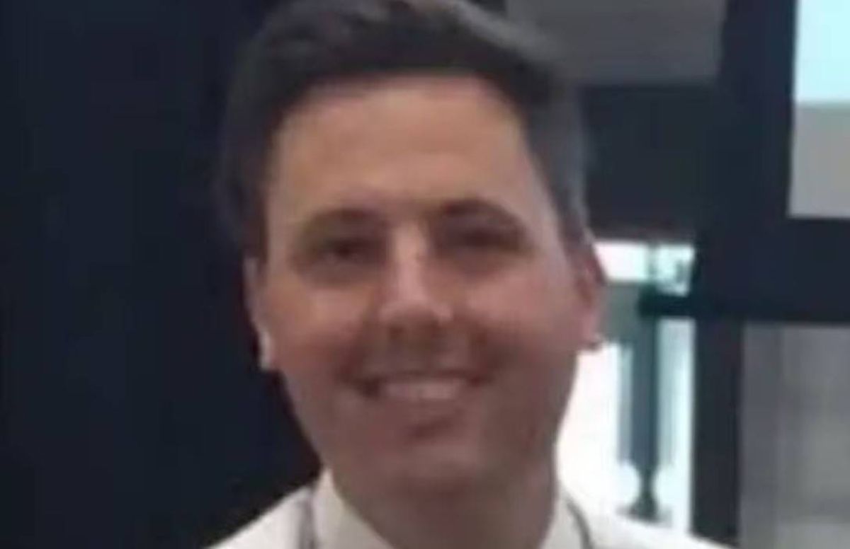 An individual with short, dark hair, dressed in a white shirt, smiling broadly while standing against a blurred background.
