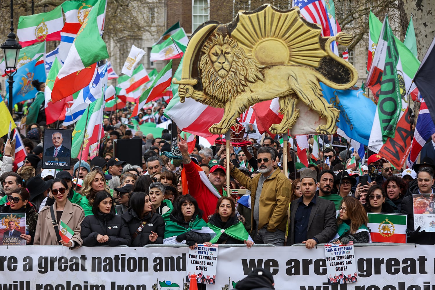 A large, crowded outdoor rally filled with people holding various flags, including versions with a lion and sun motif, alongside Israeli, British, and American flags. In the center, a man holds aloft a large, gold-colored cutout of a lion holding a sword in front of a sun. A banner at the bottom reads "we are a great nation and we will reclaim Iran."