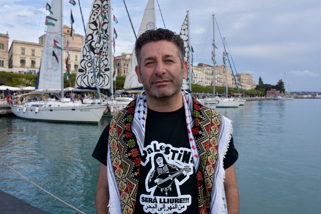Saif Abu Keshek, wearing a keffiyeh and a "Free Palestine" t-shirt, stands in front of sailboats with Palestinian flags at the Port of Siracusa.