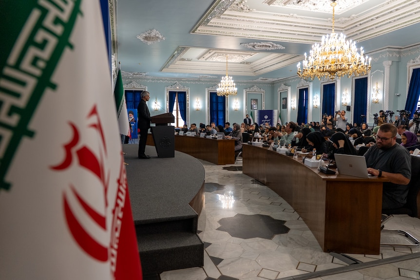 The image shows the press seated in a grandstand style series of desks and the ornate room.