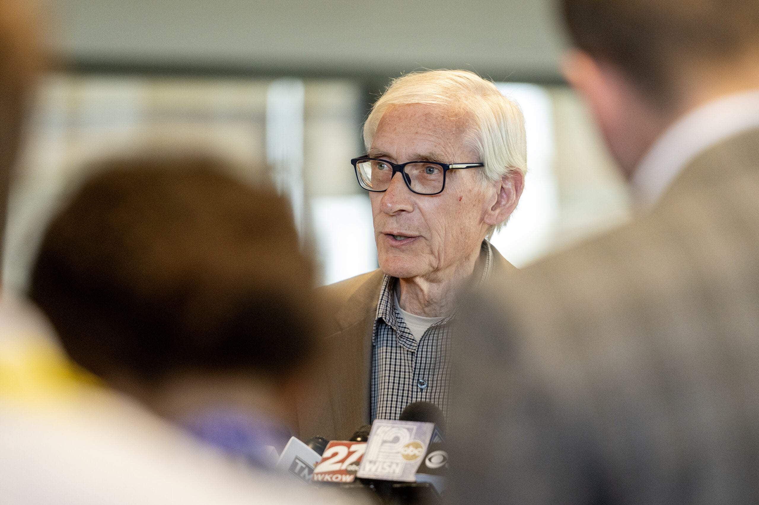 An older man with glasses speaks to reporters, microphones and news cameras in front of him, while people stand in the foreground.