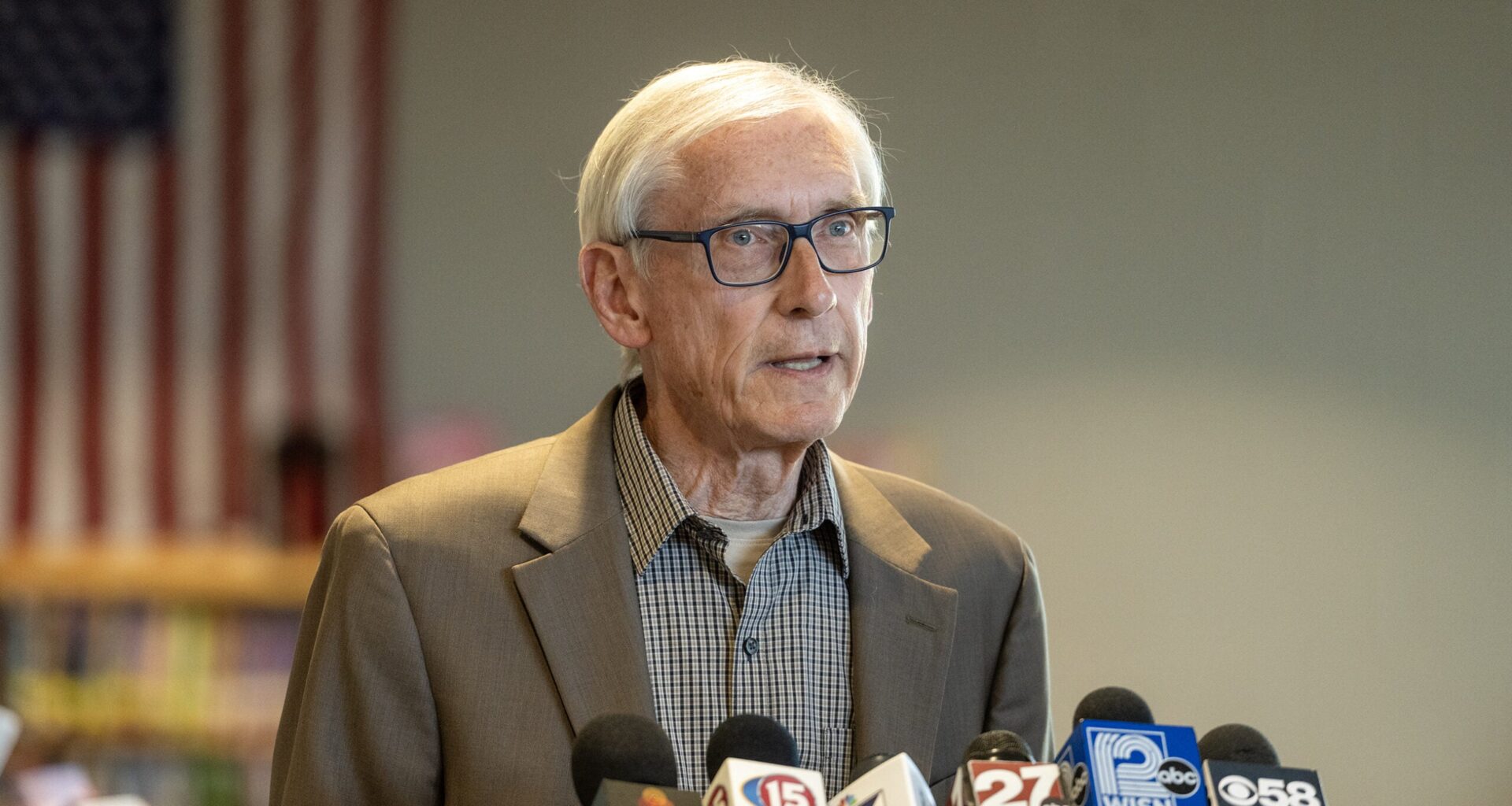 An older man in glasses and a suit speaks at a podium with several news microphones; a blurred American flag is visible in the background.