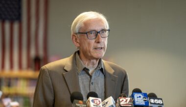 An older man in glasses and a suit speaks at a podium with several news microphones; a blurred American flag is visible in the background.
