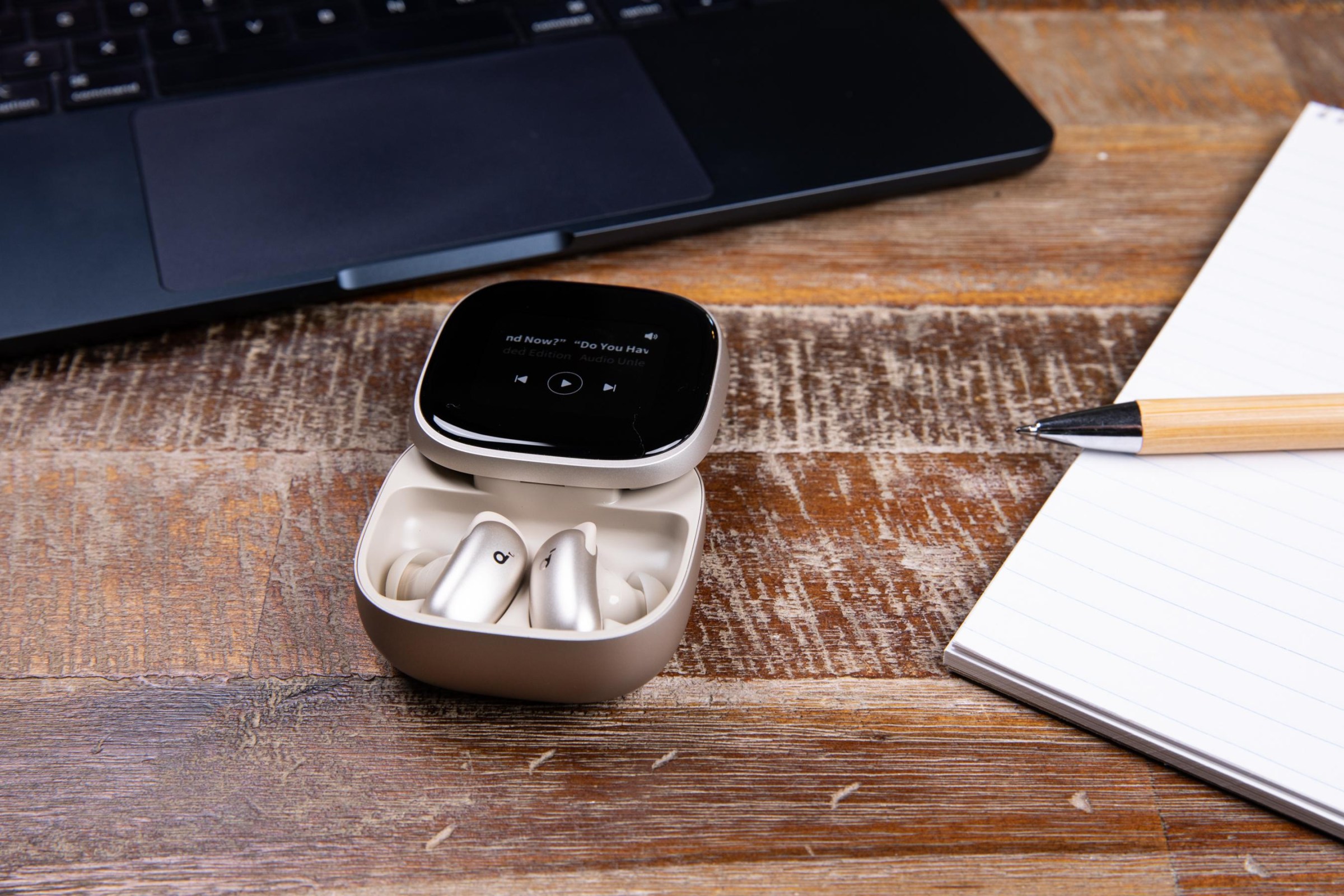 The Soundcore Liberty 5 Pro Max earbuds on a wooden coffee table next to a MacBook, pen, and paper pad.