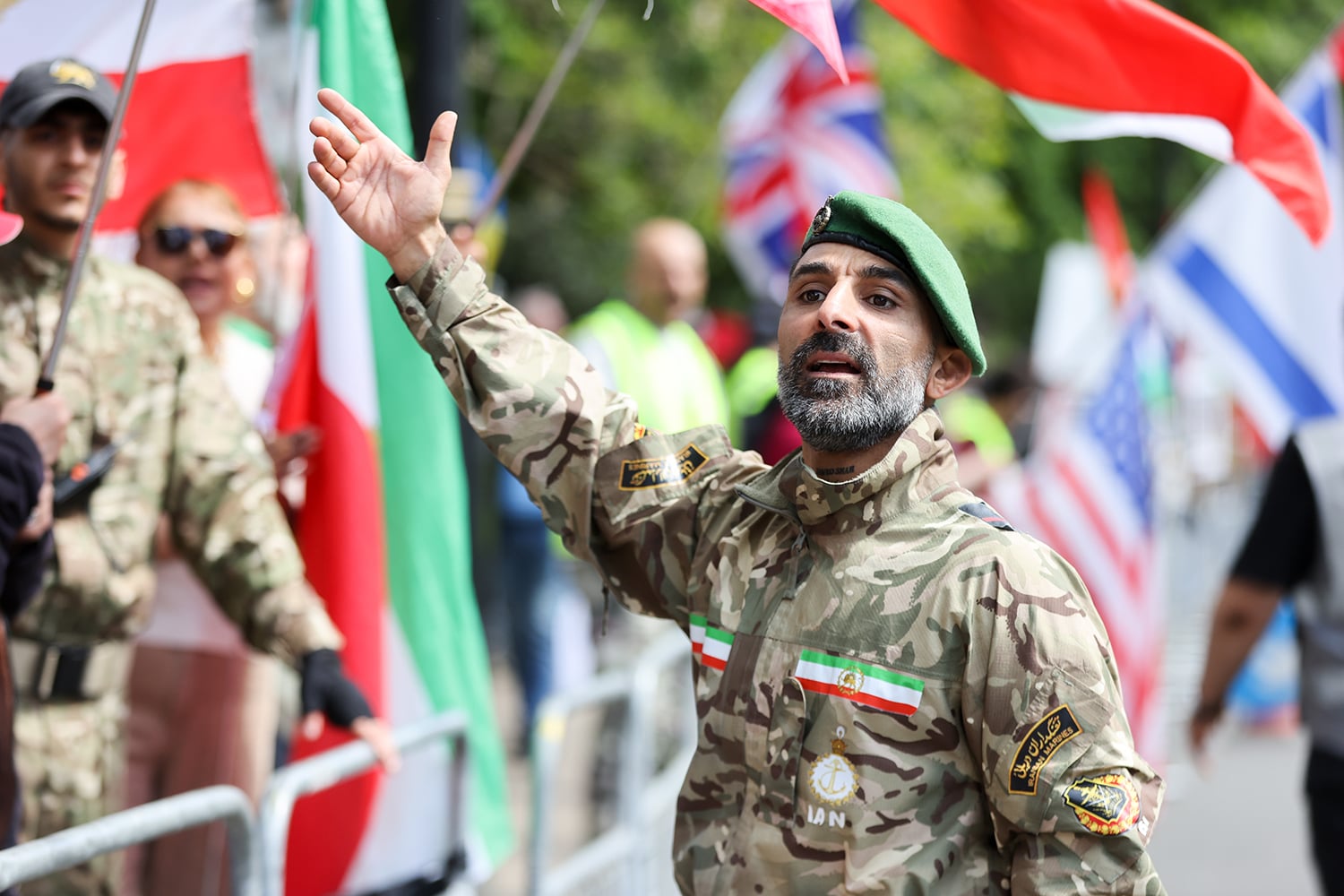 A man in a camouflage uniform and a green beret stands on a street, looking toward the camera with one arm raised. In the blurred background, other people are visible behind metal barricades holding flags, including a British flag.