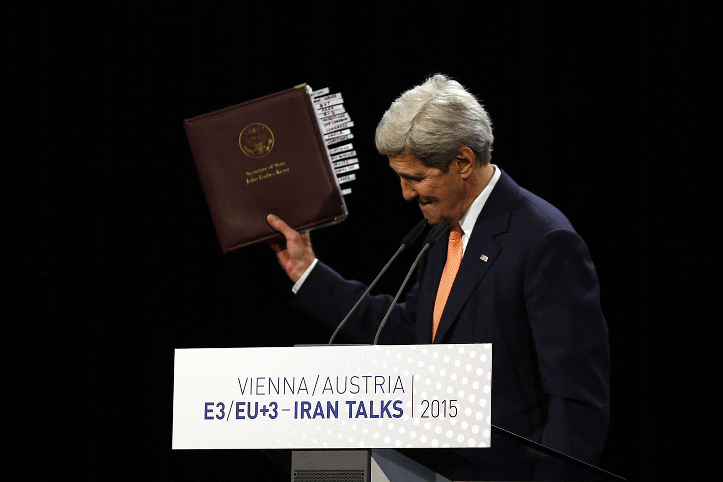 A man in a dark suit and orange tie stands behind a white podium labeled with event text. He is holding up a thick, maroon binder with many white tabs sticking out from the side. The background is solid black.